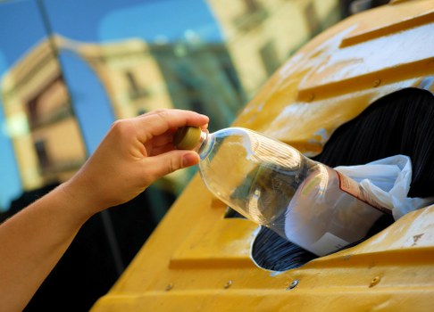 A close-up of a person's hand holding a glass jar filled with clear liquid, positioned over an open yellow rubbish bin with a black interior. The jar is partly covered with a paper or plastic lid, and the person's thumb and fingers are gripping the jar's neck. The background shows blurred outdoor environment elements with a building and blue sky, suggesting an urban or residential setting. The scene is well-lit with natural daylight, emphasizing the clean glass and the textured surface of the yellow bin, which appears to be made of plastic. The open bin is situated on a paved or concrete surface, aligned with a waste management service based in Mayfair, providing a clear visual reference for rubbish disposal or recycling activities.