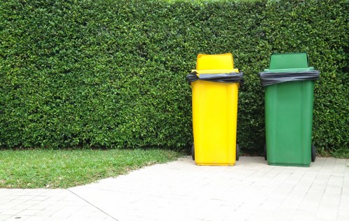 Two large wheeled rubbish bins, one yellow and one green, are positioned on a paved sidewalk in front of a dense, well-maintained hedge. The yellow bin, on the left, has a closed black lid with some black objects resting on top, while the green bin on the right has a similar black lid with no visible objects on it. The bins are made of durable plastic, with textured surfaces, and are placed side by side with a slight gap between them. The background consists of thick green foliage, indicating an outdoor urban or suburban setting, possibly near a residential or commercial property. The image suggests a rubbish collection context, which aligns with services offered by Commercial Waste Mayfair, serving the Westminster area. The lighting is even, with natural daylight illuminating the scene, and the overall environment appears tidy and orderly, typical of waste disposal zones in central London.