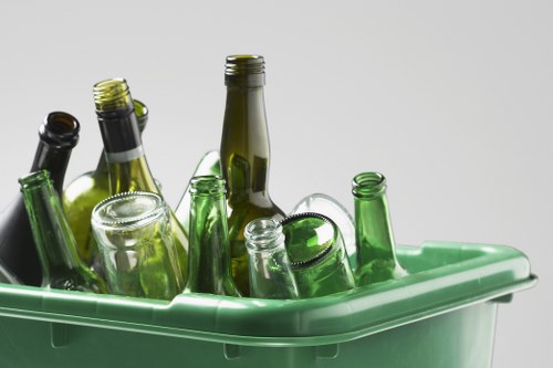 A collection of empty glass bottles, primarily green and brown, with some clear containers, partially submerged in a light green plastic recycling bin. The bottles vary in size and shape, including long-necked and shorter forms, with some caps still attached. The bin is situated on a neutral, light-coloured surface against a plain, soft grey background. The bottles are arranged with some leaning against each other, indicating they are being prepared for disposal or recycling. The green bin appears to be suitable for glass waste, commonly collected in urban waste management services in areas like Mayfair, central London. The lighting highlights the transparency and textures of the glass, capturing reflections and the smooth, glossy finishes of the bottles. This image is relevant to waste management and rubbish disposal services provided by companies such as Commercial Waste Mayfair, especially in the context of responsibly handling recyclable glass waste within the local environment.