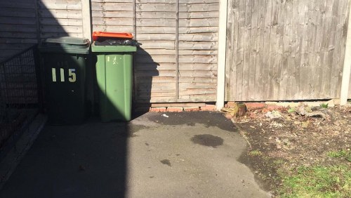 The image depicts two large wheelie bins placed on a paved outdoor surface in front of a wooden fence. One bin is dark green and the other is lighter green, both with closed lids; the green bin has the number 115 painted on its side in yellow. The bins are positioned side by side near the edge of a driveway or pavement, with the fence behind them made of vertical wooden panels. To the right, part of the fence appears weathered and slightly discolored, and there is some exposed earth and small patches of grass or weeds along the ground. The scene is lit by natural sunlight, casting shadows on the ground and fence, indicating a clear day. The image's setting suggests a typical residential area where rubbish collection or waste management services, such as those offered by Commercial Waste Mayfair, perform rubbish removal for properties in the nearby London districts, including the W1 postcode area.