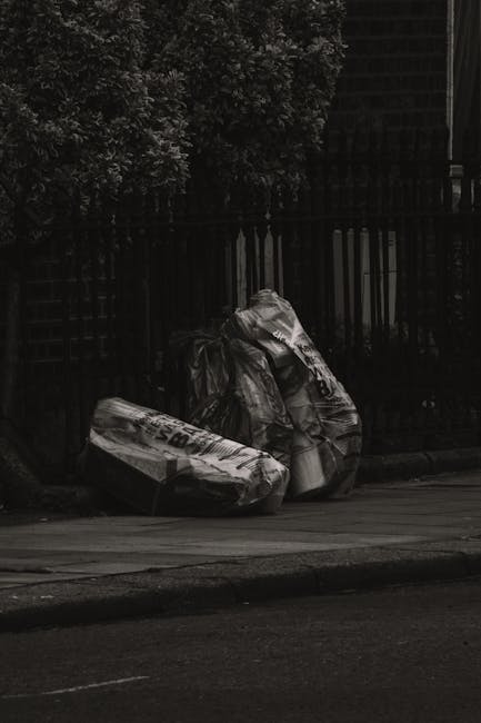 A large, green industrial rubbish skip positioned on an urban street, filled with various types of waste including crumpled paper, cardboard boxes, and discarded packaging materials. The skip shows si